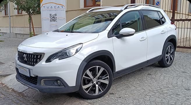 Una Peugeot 2008 di colore bianco parcheggiata su una strada in pavé, vista di tre quarti anteriore, con un'insegna 'TuaCar' visibile sullo sfondo
